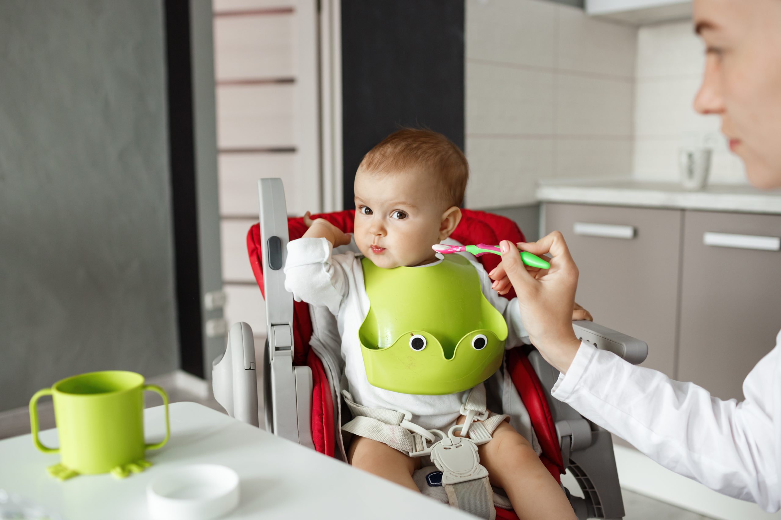 Close up of cute newborn son sitting in kitchen in baby chair and turning head aside refusing to eat